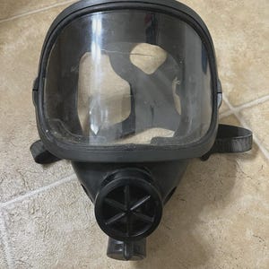 May include: A black gas mask with a clear, curved visor. The mask has a circular air intake with a star-shaped design and adjustable straps. The mask is designed to cover the entire face.