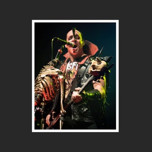 Puede incluir: Un hombre con una chaqueta de cuero negra y una camisa roja con la palabra "Misfit" está cantando en un micrófono mientras sostiene una guitarra con una calavera en el extremo. Lleva pantalones negros y botas negras.