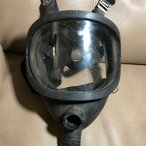 May include: A black gas mask with a clear visor and a black hose attached to the bottom. The mask has two straps that attach to the back of the head.