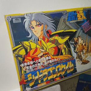 Puede incluir: Caja de juguetes de anime japonés vintage con un personaje de pelo plateado y armadura dorada. La caja tiene texto japonés y una combinación de colores dorados y rojos. También se ve una pequeña tarjeta azul.