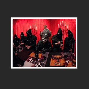 May include: A group of people in black cloaks and masks, some with hoods, are gathered around a table. One person wears a skull mask and a tall, ornate hat. The background features a red curtain and candle holders.