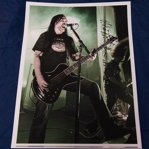 Puede incluir: Un hombre con el pelo largo y negro toca un bajo negro en el escenario. Lleva una camiseta negra con un gráfico blanco y pantalones vaqueros negros. El escenario está iluminado con luces verdes y blancas.