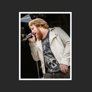 May include: A man with a beard and long hair is singing into a microphone. He is wearing a white leather jacket over a black t-shirt with a skull graphic and the text "The Anti-Christ".