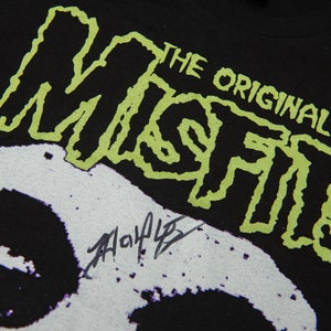 以下が含まれることがあります： 黒いTシャツに、緑と白のグラフィックで「Misfits」という単語が書かれており、その上に「The Original」というテキストがあります。Tシャツには白い骸骨のグラフィックがあり、その下に黒い署名があります。