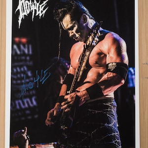 Puede incluir: Una fotografía en blanco y negro de un hombre tocando la guitarra. Lleva pantalones cortos negros con un diseño de alambre de púas y una pulsera negra con un logotipo blanco. El hombre está sin camisa y tiene el pelo negro largo. La foto está firmada "M. Shadows".