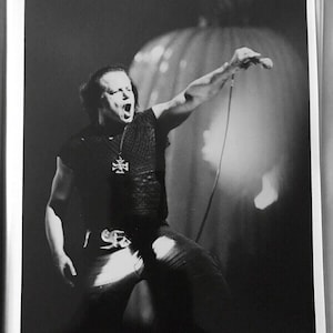 May include: Black and white photo of a person singing into a microphone. The person is wearing a black shirt with a cross pendant and black pants. The person is on stage with a spotlight behind them.