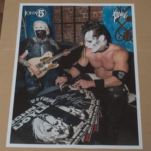 May include: A promotional poster featuring two musicians, one playing a guitar and the other signing autographs. The poster includes the text "JOHN 5" and "MAD MONSTER TOUR". The background features a brick wall.