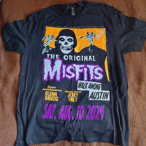 May include: Black t-shirt featuring a graphic of a skull with the text "THE ORIGINAL MISFITS" in purple and white. The shirt also includes the text "WALK AMONG AUSTIN" and the date "SAT. AUG. 10 2024".