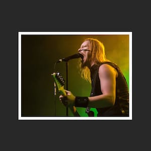 May include: A man with long blonde hair sings into a microphone while playing a green electric guitar. He is wearing a black sleeveless shirt and has black tape across his face. The background is a dark green and yellow.