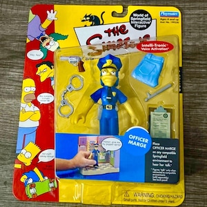 May include: A packaged Officer Marge action figure from The Simpsons. The figure is in a blue police uniform with accessories including handcuffs, a gun, and a clipboard. The packaging includes the text "World of Springfield Interactive Figure."