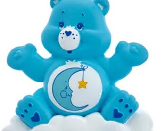 May include: A light blue Care Bear ornament. The bear is sitting on a white cloud and has a crescent moon and star design on its belly. The ornament has a silver chain for hanging.