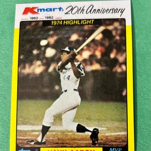 May include: A baseball card featuring Hank Aaron in action, swinging a bat. The card is a Kmart 20th Anniversary edition from 1974, with the Topps logo. The card has a yellow border and includes the text "MVP Series".