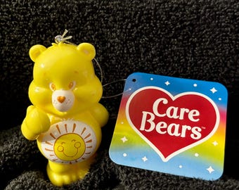 May include: A yellow Care Bear ornament with a smiling sun design on its belly. The ornament has a silver string for hanging and a tag with the Care Bears logo in a heart shape.