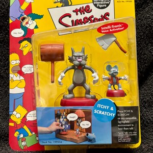 May include: A Playmates World of Springfield Interactive Figure set featuring the characters Itchy and Scratchy. The packaging is yellow and red, with images of The Simpsons characters. The set includes a mallet, axe, and a small stage.