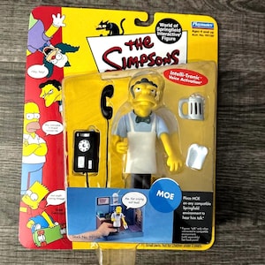 May include: A packaged "World of Springfield" interactive figure of Moe from The Simpsons. The figure is yellow with a white apron, blue shirt, and black hair. Accessories include a beer mug, napkin, and telephone. The packaging features other Simpsons characters.