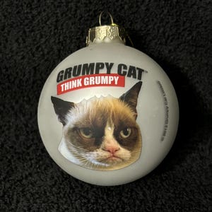 May include: A round, gray glass Christmas ornament with a picture of Grumpy Cat on it. The ornament has the text "Grumpy Cat" and "Think Grumpy" printed on it.