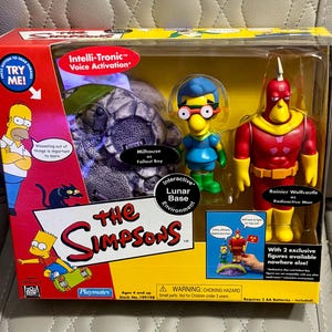 May include: A The Simpsons Playmates toy set in a rectangular box featuring Milhouse as Fallout Boy and Rainier Wolfcastle as Radioactive Man figures, along with a lunar base environment. The packaging includes text, graphics, and a try-me button.