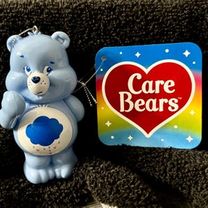 May include: A light blue Care Bear figurine with a cloud belly badge and a sad expression. The figurine is holding up its right hand. A tag with the Care Bears logo is attached.