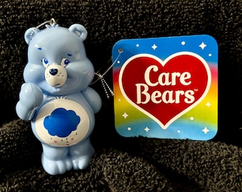 May include: A light blue Care Bear figurine with a cloud belly badge and a sad expression. The figurine is holding up its right hand. A tag with the Care Bears logo is attached.