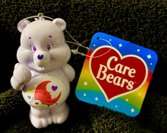 May include: A lavender Care Bear ornament with purple accents, including the eyes and nose. The bear has a crescent moon and heart design on its belly. A tag with the Care Bears logo is attached.