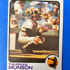 May include: A baseball card featuring Thurman Munson of the New York Yankees. Munson is shown in his catcher's gear, crouched down. The card has a white border and includes the team name and position.