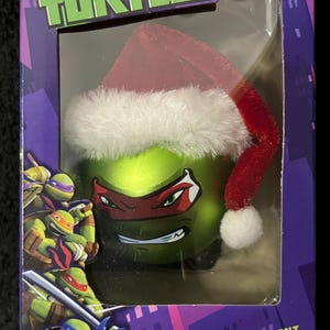 May include: A Teenage Mutant Ninja Turtles Christmas ornament featuring a green, red, and white Santa hat on a green turtle head with a red mask. The ornament is in a box with a purple background and the words "Teenage Mutant Ninja Turtles" and "Christmas Ornament".