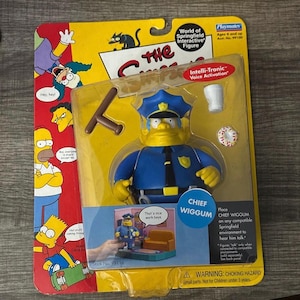 May include: A yellow plastic Chief Wiggum action figure from The Simpsons. The figure is wearing a blue police uniform with a badge that says "Chief Wiggum". The figure is holding a nightstick in one hand and a donut in the other. The figure is standing on a small platform with a scene from the Simpsons cartoon in the background. The packaging says "Intelli-Tronic Voice Activation" and "World of Springfield Interactive Figure".