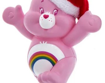 May include: Pink Care Bear Christmas ornament wearing a red and white Santa hat. The bear has a rainbow on its belly and is posed with its arms outstretched. A festive holiday decoration.