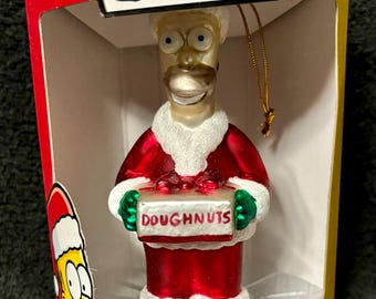 May include: A glass Christmas ornament featuring Homer Simpson dressed as Santa Claus holding a box of donuts. The ornament is packaged in a yellow box with the words "The Simpsons" on the top and "Hand-Crafted Glass Holiday Ornament" on the bottom.