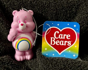 May include: A pink Care Bear ornament with a rainbow belly and a heart-shaped nose. The ornament has a silver string for hanging and is next to a tag with the words "Care Bears" in a heart shape.
