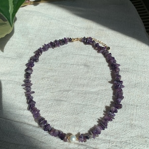 May include: A purple amethyst stone necklace with a single white pearl accent. The necklace is made of small, irregular shaped amethyst stones and is strung on a gold chain.