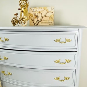 May include: A light gray dresser with four drawers and gold-toned hardware. The dresser has a curved front and a slightly rounded top. The dresser is decorated with a gold-toned bicycle figurine and a framed print with the text "family is..."