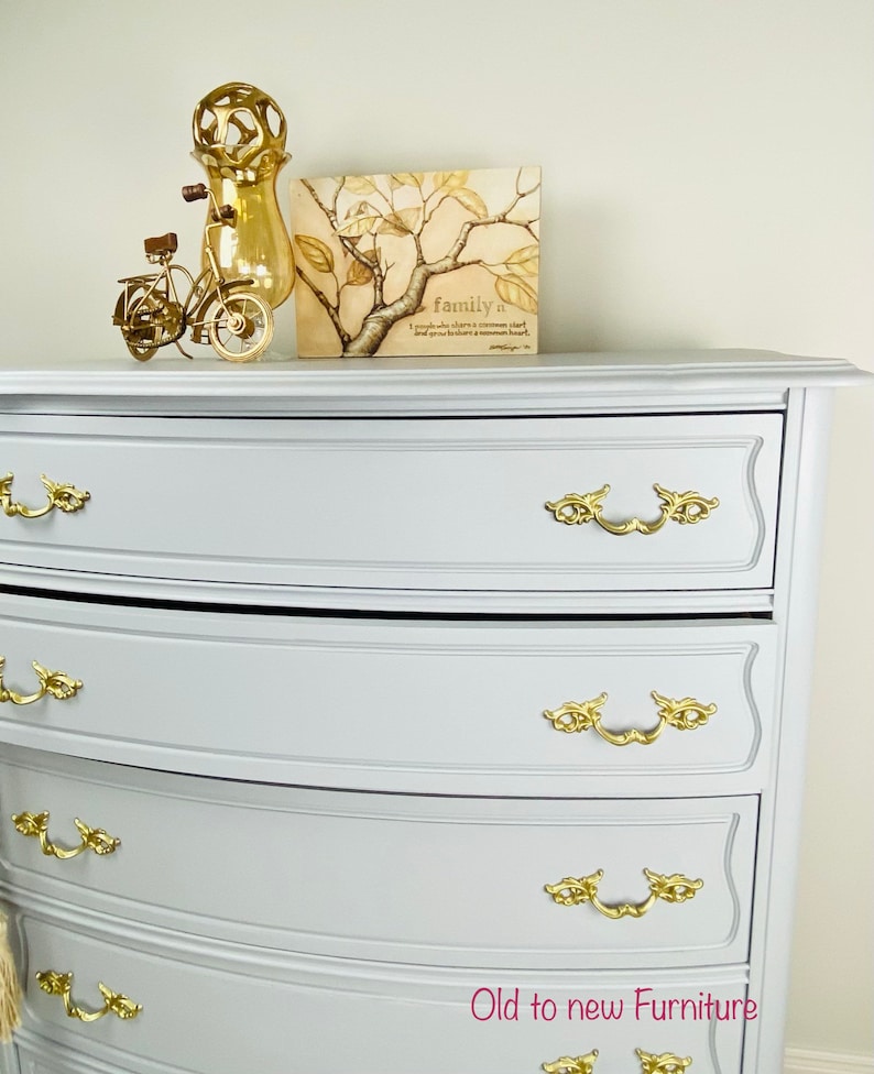 May include: A light gray dresser with four drawers and gold-toned hardware. The dresser has a curved front and a slightly rounded top. The dresser is decorated with a gold-toned bicycle figurine and a framed print with the text "family is..."