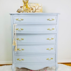 May include: A light blue dresser with six drawers and gold-toned hardware. The dresser has a curved front and sits on four legs. The dresser is decorated with a gold-toned decorative object and a framed print that says "Family".
