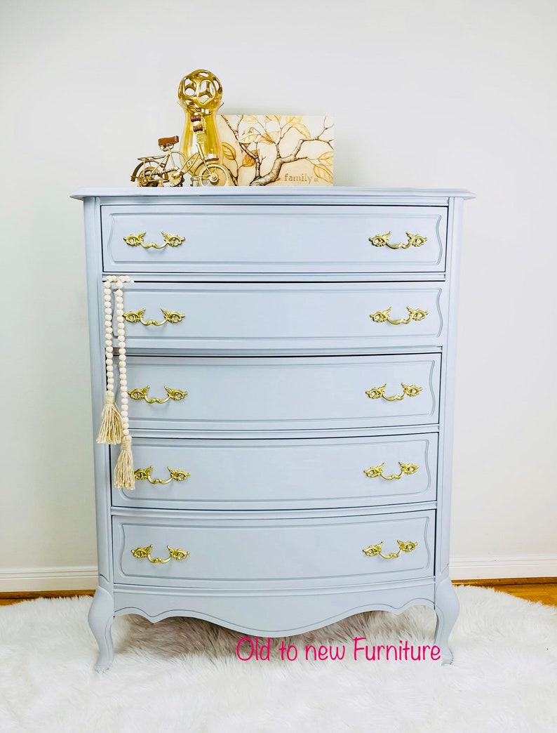 May include: A light blue dresser with six drawers and gold-toned hardware. The dresser has a curved front and sits on four legs. The dresser is decorated with a gold-toned decorative object and a framed print that says "Family".