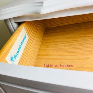 May include: A white painted wooden dresser drawer with a gold drawer pull. The drawer is open and the inside is made of wood. A blue and white label with the text "Broyhill Premier" is attached to the inside of the drawer. The text "Old to new Furniture" is written in pink on the bottom of the drawer.