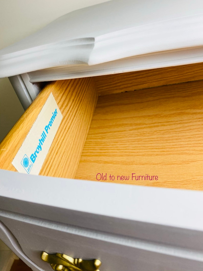 May include: A white painted wooden dresser drawer with a gold drawer pull. The drawer is open and the inside is made of wood. A blue and white label with the text "Broyhill Premier" is attached to the inside of the drawer. The text "Old to new Furniture" is written in pink on the bottom of the drawer.