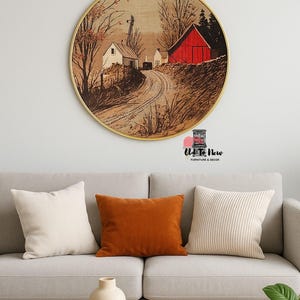 May include: A round wall art piece with a rural landscape scene, featuring a red barn and white buildings, hangs above a gray sofa with orange and cream pillows. A wooden coffee table with a vase and books sits in front.