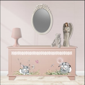 Puede incluir: Un cofre de madera rosa con un borde de encaje blanco y un diseño caprichoso con dos conejos y dientes de león. El cofre está decorado con una lámpara rosa claro, un oso de peluche gris y una figura de ángel blanco.