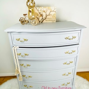 May include: A gray painted dresser with five drawers and gold-toned hardware. The dresser has a curved top and bottom and sits on a white fluffy rug. The dresser is decorated with a gold metal vase, a small gold bicycle, and a framed print with the text "Family is a circle of people who love and support each other. It is a place where you feel safe and loved. It is a place where you can be yourself."