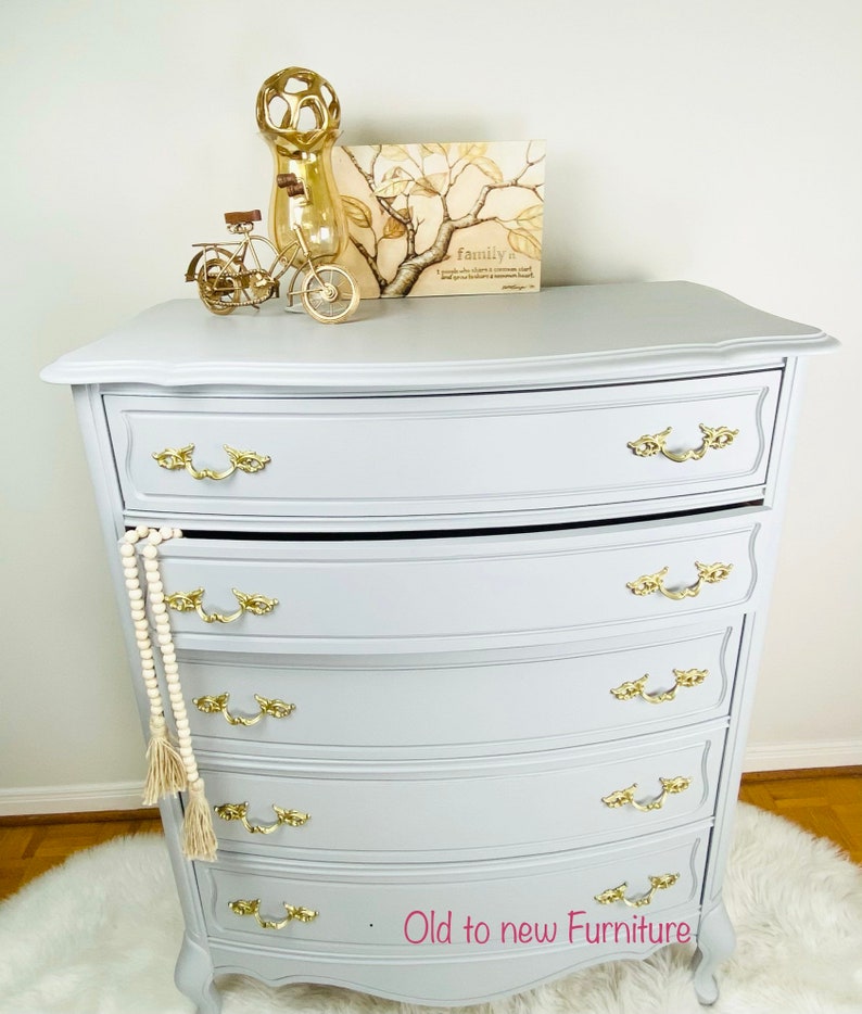 May include: A gray painted dresser with five drawers and gold-toned hardware. The dresser has a curved top and bottom and sits on a white fluffy rug. The dresser is decorated with a gold metal vase, a small gold bicycle, and a framed print with the text "Family is a circle of people who love and support each other. It is a place where you feel safe and loved. It is a place where you can be yourself."