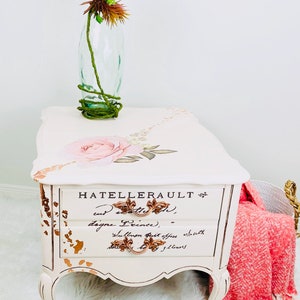May include: White painted end table with gold accents and floral design. The table has two drawers with gold handles and the text "HATELERERAULT" is written on the front. The table is sitting on a white fluffy rug.