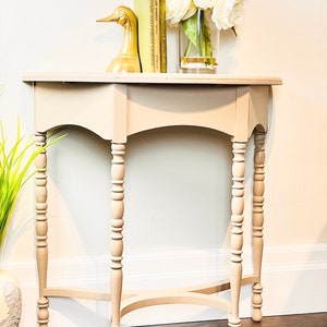 May include: A light beige, half-moon shaped side table with turned legs and a decorative X-shaped stretcher. The table is decorated with a gold duck figurine and a vase of white flowers.