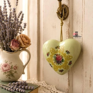 May include: A pale green ceramic heart ornament hanging from a brass hook by a gold cord. The heart features a central yellow filigree frame surrounding a floral bouquet of pink and purple roses, with smaller floral accents scattered across the surface.