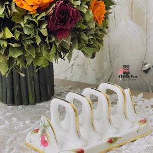 Puede incluir: Un tostador de cerámica blanca con ribetes dorados y detalles florales. El soporte tiene cuatro ranuras arqueadas y está decorado con flores rojas y rosas. Un ramo de rosas naranjas y burdeos se encuentra en el fondo.