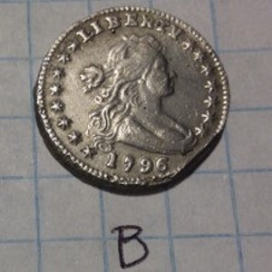 May include: A silver coin with a profile of a woman, the word "LIBERTY" above, and the date "1996" below. The coin has a star pattern around the edge and the letter "B" is written below the coin.