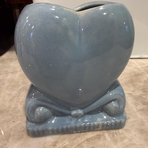 May include: A pale blue ceramic heart-shaped vase. The vase has a decorative base with scroll details and a small opening at the top. The vase has a glossy finish and is a solid colour.