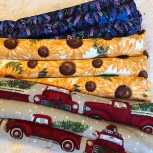 Op de afbeelding: Vier stoffen warmtepads, elk met een ander ontwerp. Het bovenste pad heeft een paars bloemenpatroon op een zwarte achtergrond. Het tweede pad heeft een geel zonnebloemmotief op een witte achtergrond. Het derde pad heeft een grijze achtergrond met witte stippen en een rode vrachtwagen met een kerstboom op de laadbak. Het onderste pad heeft een vergelijkbaar ontwerp als het derde, maar de vrachtwagen is in de tegenovergestelde richting gericht.
