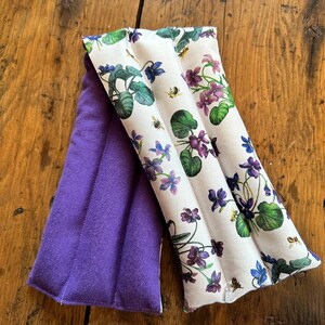 Op de afbeelding: Een paarse fleece gevoerde nekwarmer met een witte stoffen inzet met een bloemenpatroon van paarse viooltjes en groene bladeren. De stoffen inzet heeft een herhalend patroon van bijen.