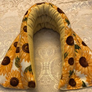 Op de afbeelding: Een gele nekwarmer met een zonnebloemprint. De nekwarmer is gemaakt van zachte stof en is gevuld met lijnzaad. Hij kan worden verwarmd in de magnetron of gekoeld in de vriezer om pijn en spanning te verlichten.